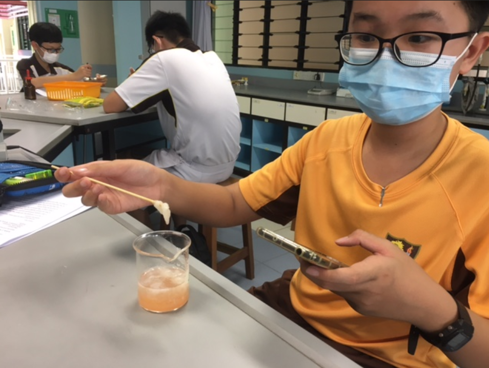 Student in mask and glasses holds a wooden stick with white substance over a small beaker of pink liquid in a lab.